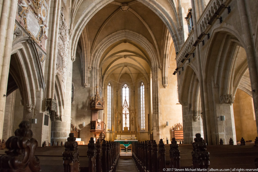 Gothic church nave with ribbed vaults, stone piers, dark pews, and tall sanctuary windows above the altar.