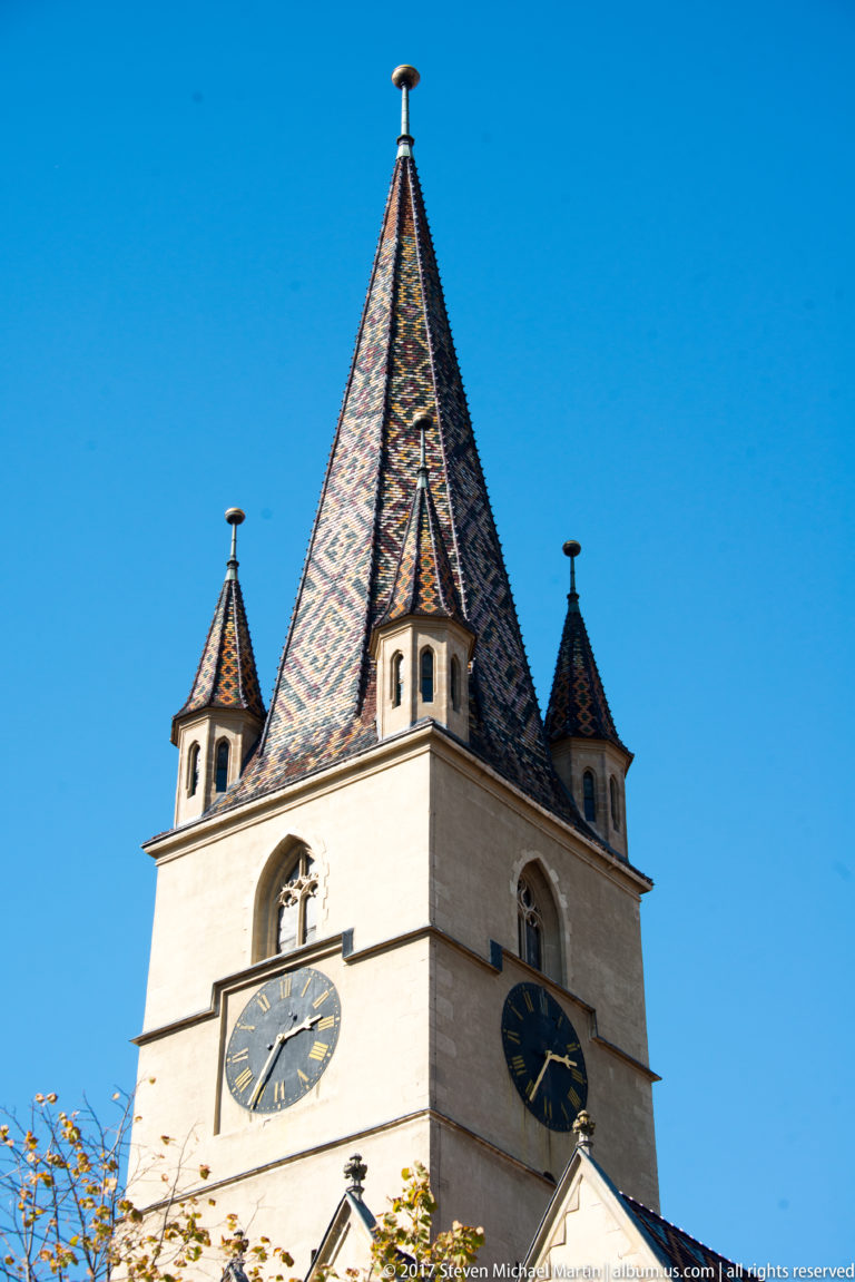 Pale church tower with two clock faces, a steep multicolored tiled roof, and small corner turrets against blue sky.
