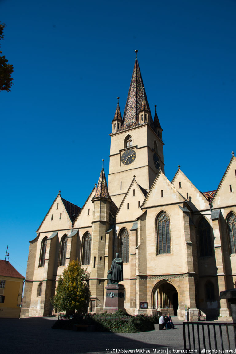 Large Gothic church with tall clock tower, buttressed walls, pointed windows, and a statue in the paved forecourt.