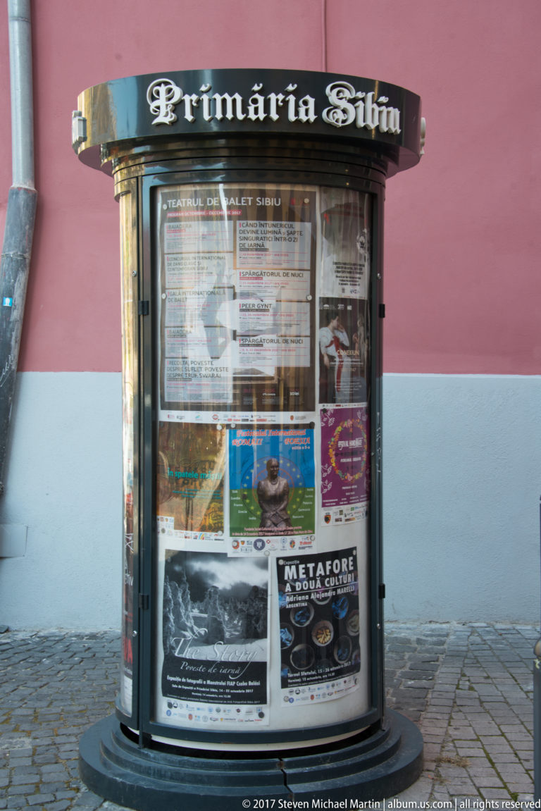Cylindrical Primăria Sibiu information kiosk filled with theater, concert, and event posters beside a pink wall.