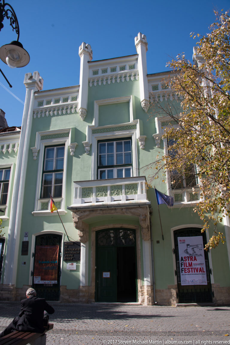 Pale green historic cultural building with white ornament, balcony, flags, and Astra Film Festival posters at the entrance.