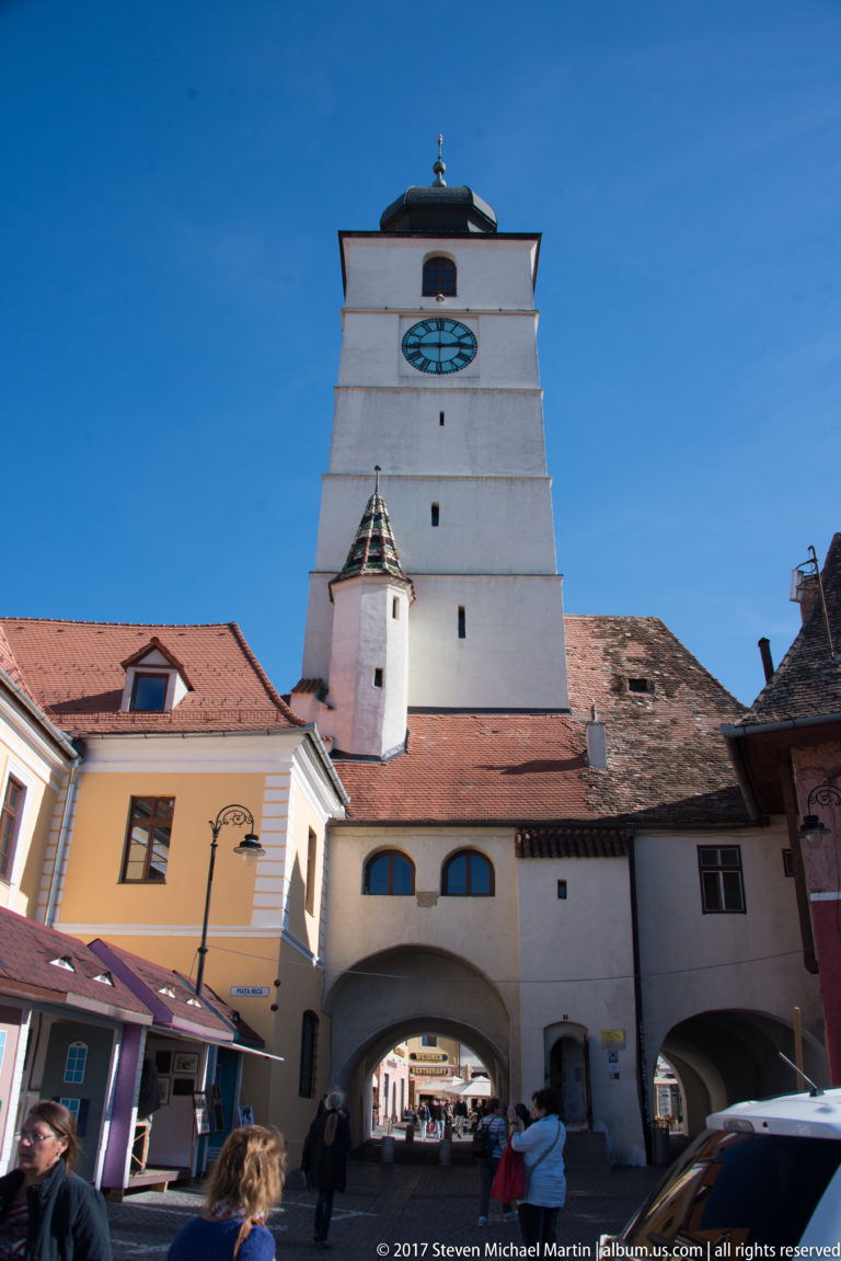 White clock tower above a broad arched street gateway, with pedestrians passing through the vaulted opening below.