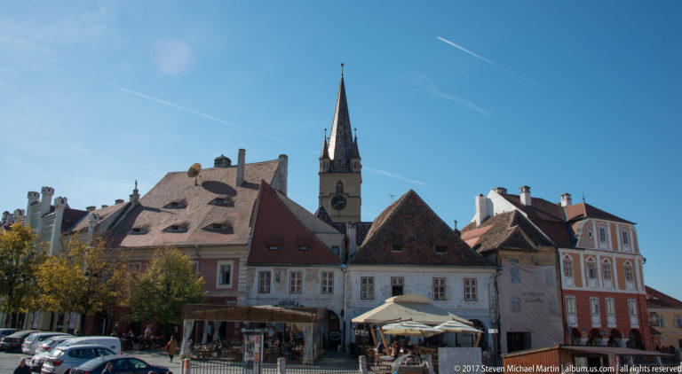 Historic Sibiu square lined by pastel façades and steep roofs, with café seating and a slender church spire behind.