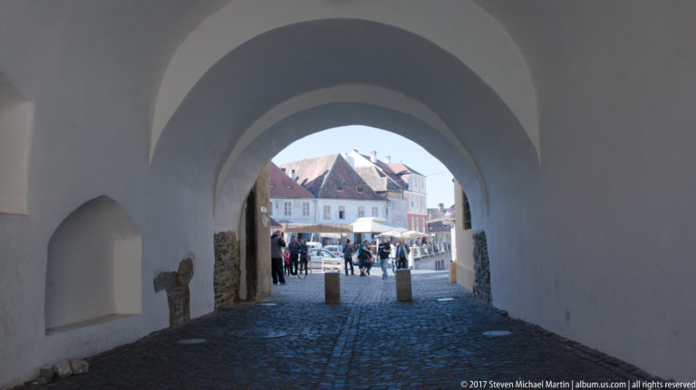 White vaulted pedestrian passage opening from shade onto a sunlit cobbled square with people and café umbrellas.