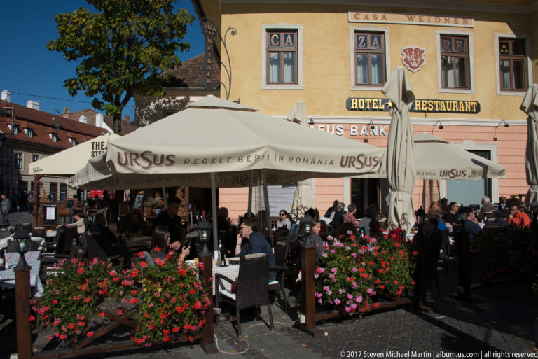 Crowded outdoor café beneath Ursus umbrellas and Casa Weidner hotel-restaurant signage, with bright flower planters in front.