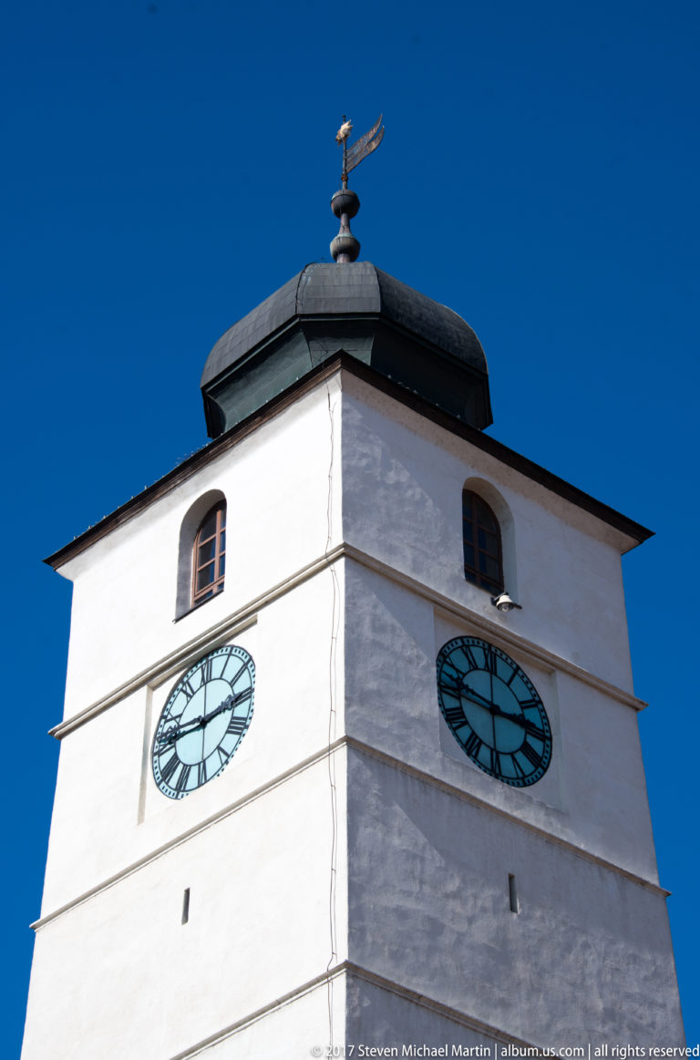 White square tower with two pale-blue clock faces, small openings, and a broad dark roof against blue sky.