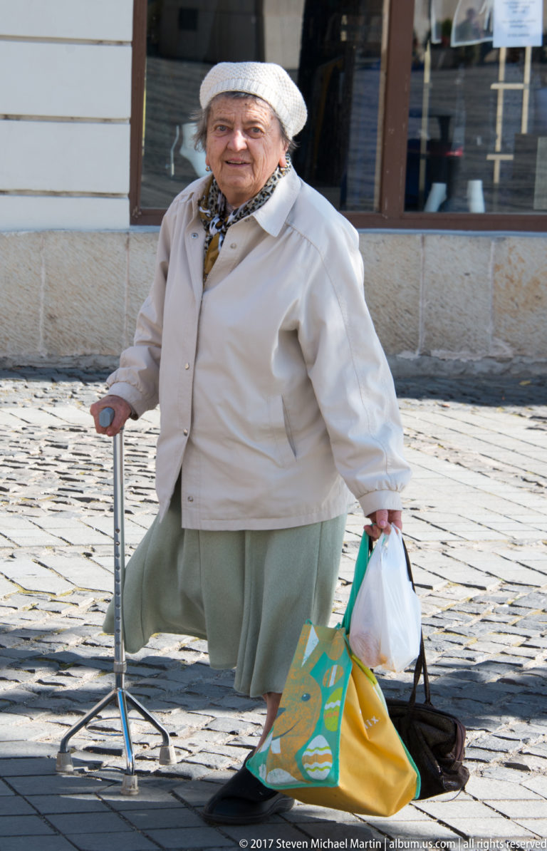 Elderly woman in a light coat and knit hat using a cane while carrying shopping bags across cobblestones.