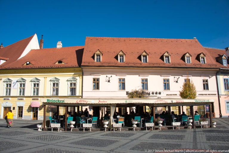 Long pale historic building with red-tiled dormer roof and a row of cafés and outdoor tables along the ground floor.
