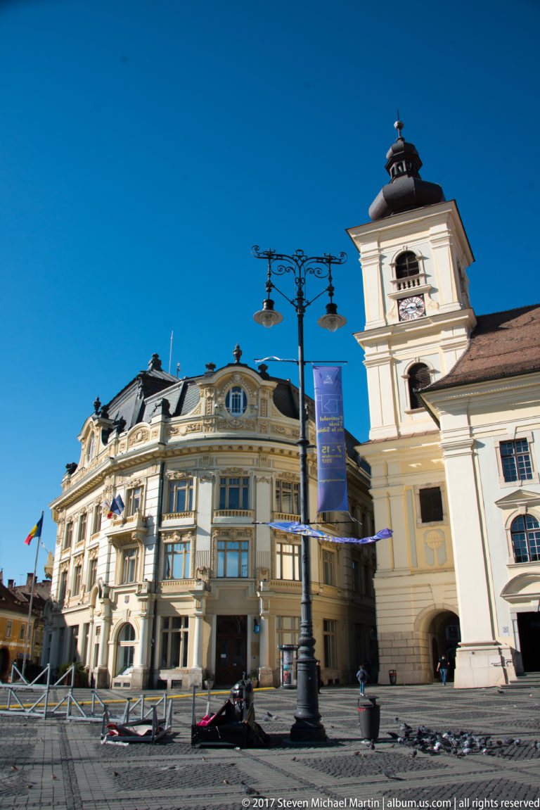 Ornate cream civic building beside a church tower on a cobbled Sibiu square, with a decorative streetlamp in front.