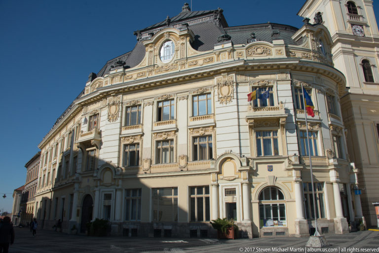 Ornate historic corner building with carved façade, Romanian and EU flags, and a clock tower rising at the far side.