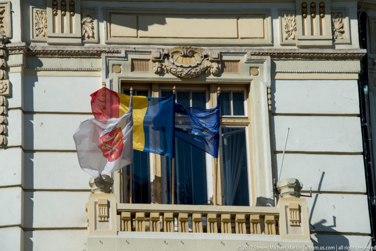 Romanian, European Union, and municipal flags hanging beside a tall window on an ornate historic façade.