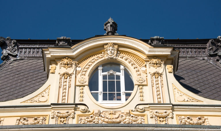 Curved ornate pediment with oval window, sculpted faces, scrollwork, garlands, and dark slate roofing.