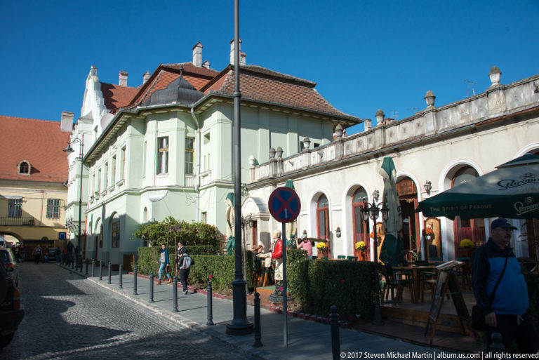 Narrow cobbled Sibiu street with pastel buildings, red roofs, pedestrians, planters, and outdoor restaurant seating.