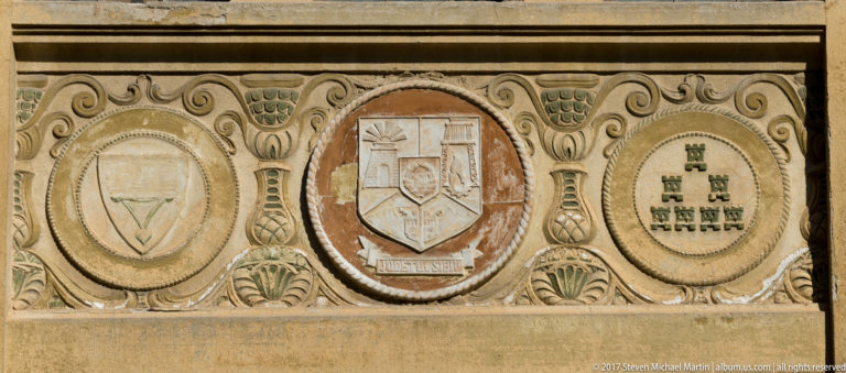 Weathered carved stone panel with three heraldic shields set in oval medallions and surrounded by scrolling ornament.