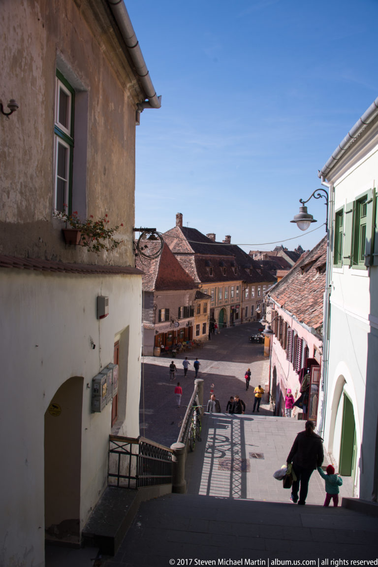 Shaded passage overlooking a small Sibiu square with pedestrians, weathered façades, and red roofs.