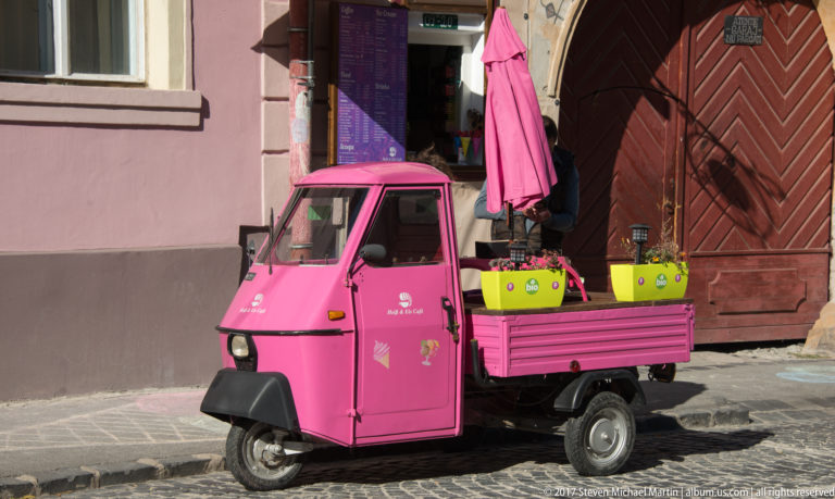 Bright pink three-wheeled utility vehicle parked by a wooden doorway, carrying colorful flower planters and a folded umbrella.