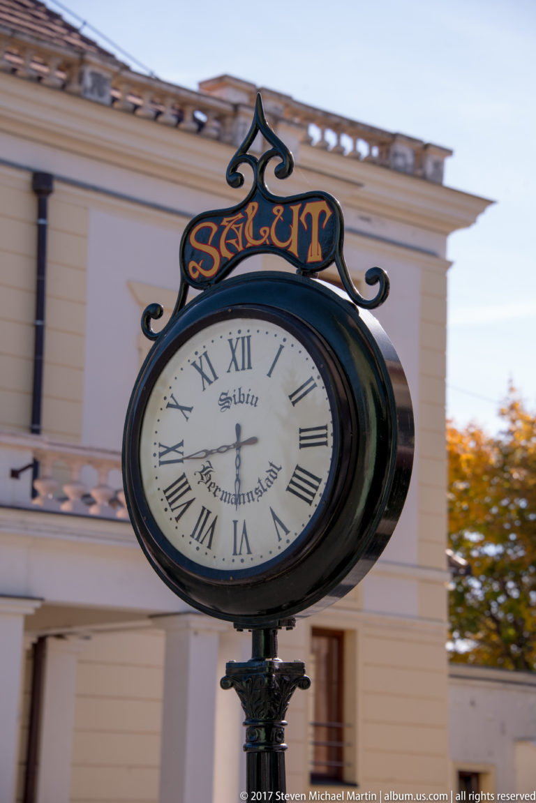 Ornamental black street clock with Roman numerals and the words Sibiu and Hermannstadt on its white face.