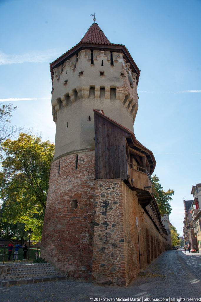 Round defensive tower with projecting upper stage, red-tiled roof, brick wall, and covered wooden gallery in Sibiu.