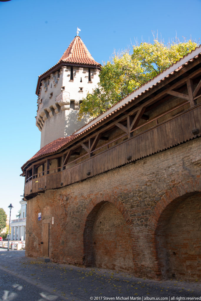 Long Sibiu brick fortification wall with blind arches, covered timber gallery, and a tiled-roof defensive tower at one end.
