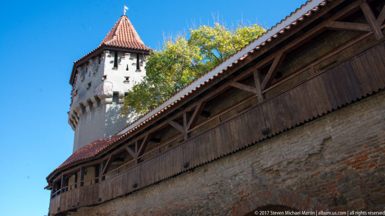 Low-angle view of a wooden covered gallery atop a brick wall with a pale defensive tower rising behind.