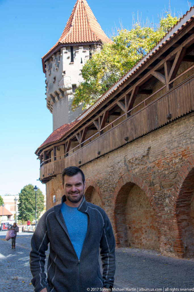 Man standing before a long brick fortification wall and tiled-roof defensive tower in Sibiu.