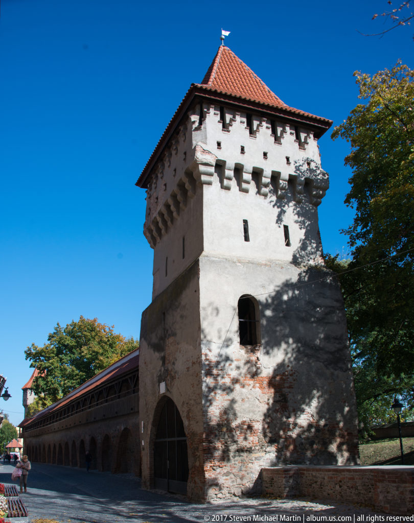 Square defensive tower with corbelled upper story, slit openings, red-tiled roof, and long covered wall extending beside it.
