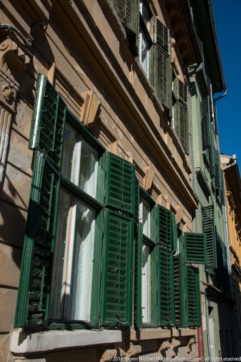 Historic façade with many dark green louvered shutters opened around white-framed windows, seen from a steep upward angle.