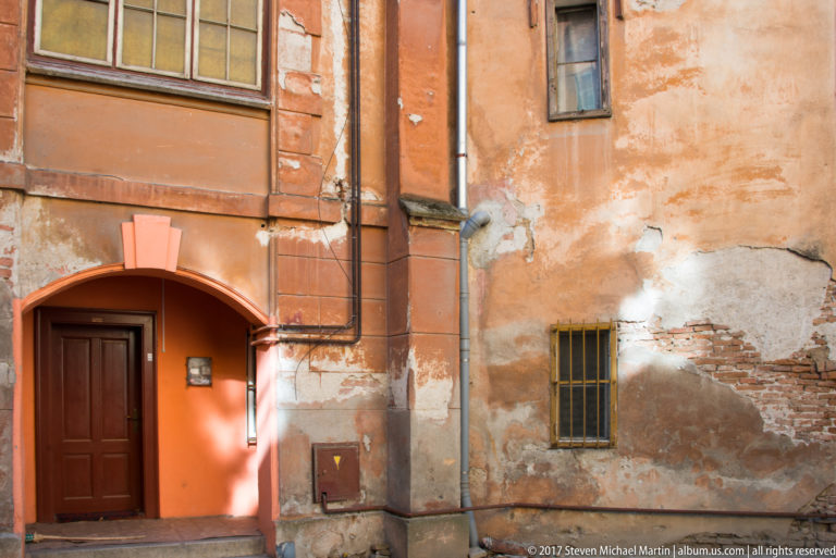 Weathered orange wall with dark wooden door, barred window, exposed brick, peeling plaster, pipes, and utility boxes.