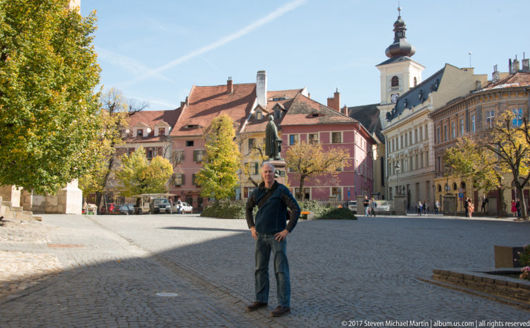 Large cobbled square in Sibiu with a standing person, historic façades, autumn trees, and a white church tower.