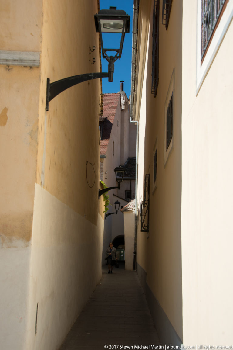Narrow shaded pedestrian alley between pale walls, with black hanging lanterns leading toward a brighter opening in the distance.