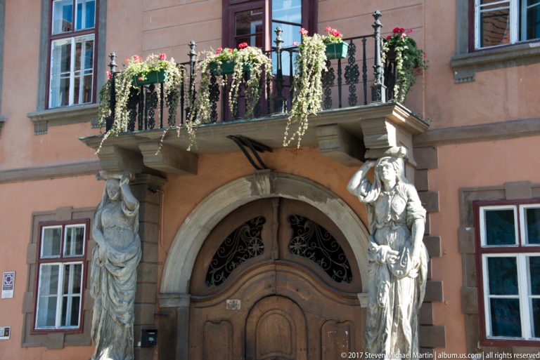 Historic Sibiu doorway with two caryatid-like figures supporting a wrought-iron balcony filled with cascading flowers.