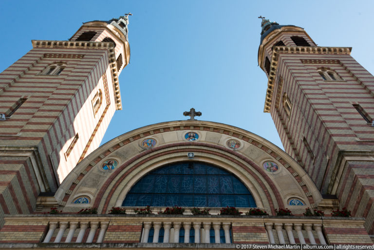 Low-angle view of Holy Trinity Orthodox Cathedral with twin striped towers, arched central window, decorative roundels, and cross.
