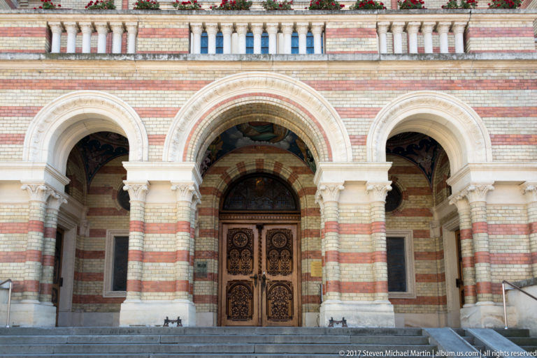 Triple-arched cathedral entrance with striped masonry, carved wooden doors, columns, balustrade, and flowers.