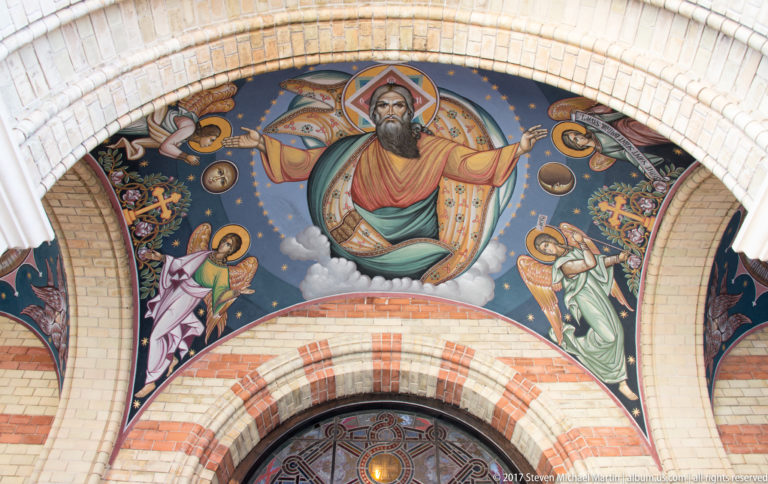 Exterior Orthodox mosaic of Christ blessing, surrounded by angels and celestial motifs above an arched cathedral doorway.
