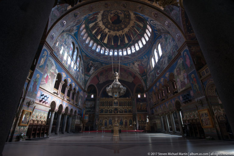 Wide interior of Holy Trinity Orthodox Cathedral with frescoed dome, chandelier, painted walls, choir stalls, and gilded iconostasis.