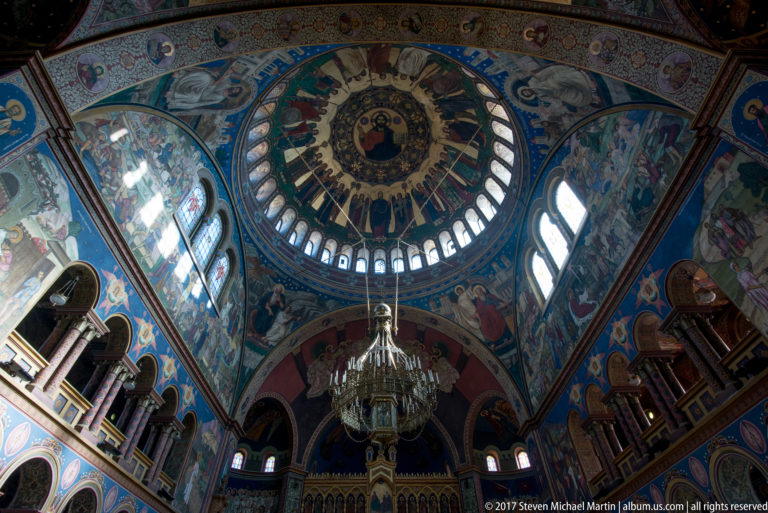 Upward view of a richly frescoed Orthodox dome with ring windows, biblical scenes, saints, and a large hanging chandelier.