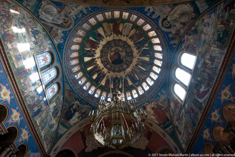 Direct upward view of Orthodox cathedral dome with central sacred image, ring windows, radiating frescoes, and chandelier.