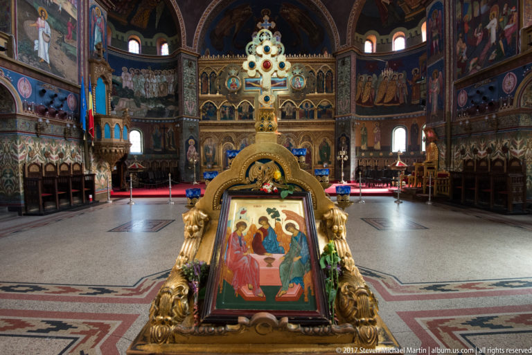 Framed Orthodox icon on an ornate gilded stand with flowers, positioned before the cathedral iconostasis.
