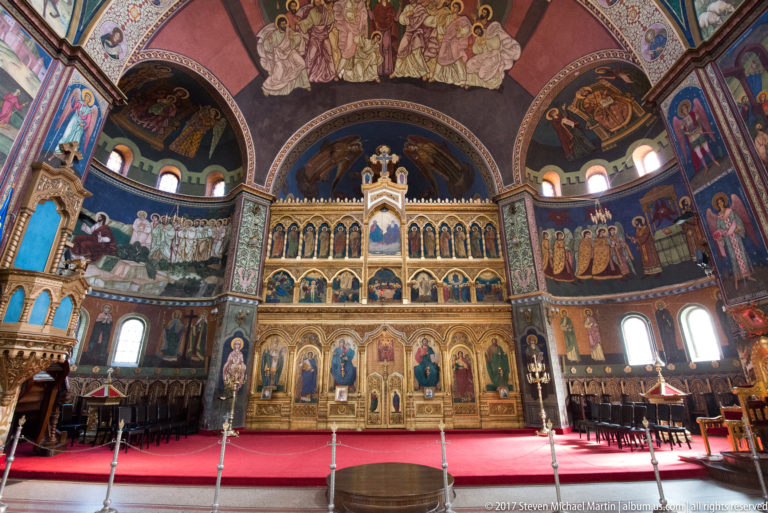Frontal view of gilded Orthodox iconostasis beneath extensive frescoes, with red carpet and choir furnishings.