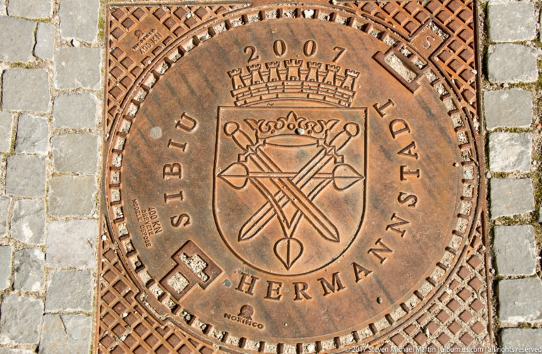 Round metal manhole cover reading Sibiu Hermannstadt and 2007, with a civic crest set in stone paving.