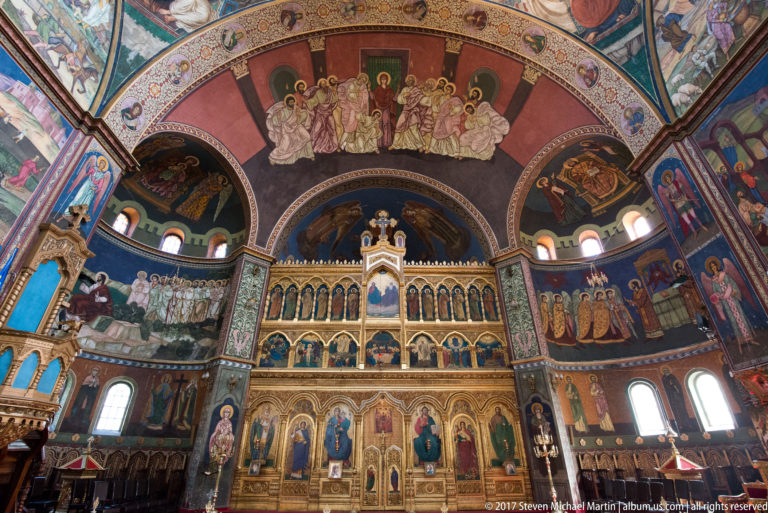 Upper cathedral sanctuary with gilded iconostasis, rows of icons, large narrative frescoes, arches, and high windows.