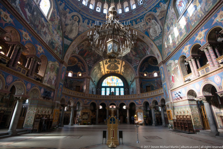Rear-facing cathedral nave with large chandelier, frescoed vaults, arcaded galleries, wooden stalls, and bright end window.