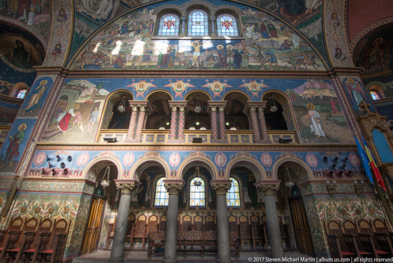 Cathedral side wall with two-level rounded arcade, striped columns, wooden choir stalls, frescoes, and high windows.