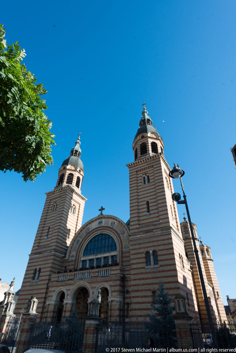 Street-level view of Holy Trinity Orthodox Cathedral with twin towers, striped masonry, central arch, and triple entrance.