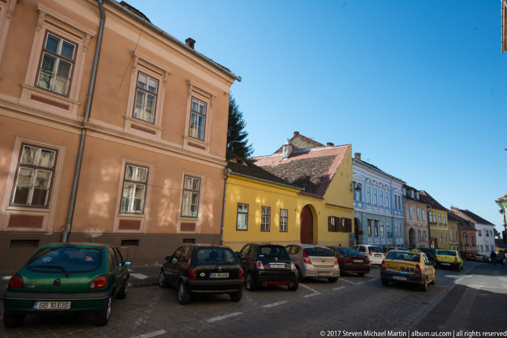 Pastel historic houses with tiled roofs and many parked cars along a quiet street in Sibiu.