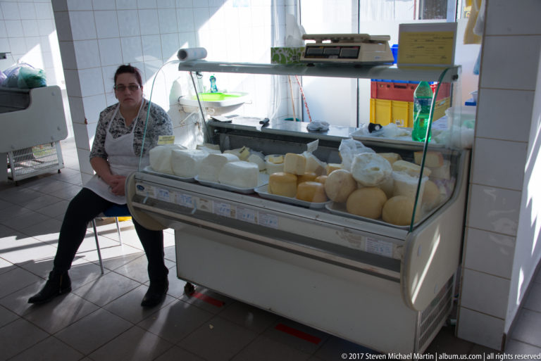 Seated market vendor beside a refrigerated glass case containing rounds and blocks of pale cheese.