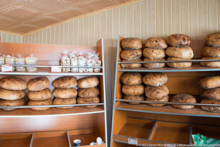 Wooden market shelves stacked with large flour-dusted round and oval rustic bread loaves.