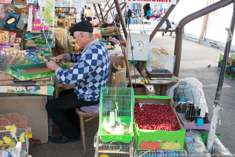 Seated vendor at a crowded market stall with red produce, packaged goods, and cages containing small birds.