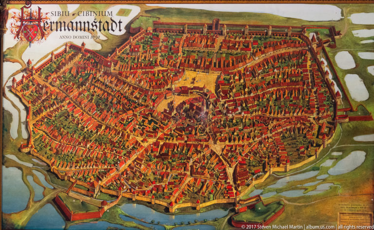 Illustrated bird’s-eye plan of fortified Sibiu with walls, towers, dense red roofs, and surrounding waterways.