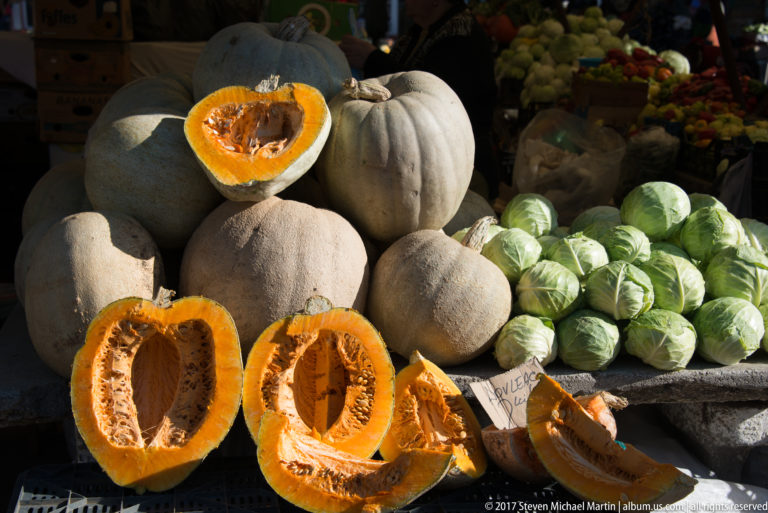 Cut orange pumpkins with seeds beside whole gray pumpkins and pale green cabbages at a market stall.